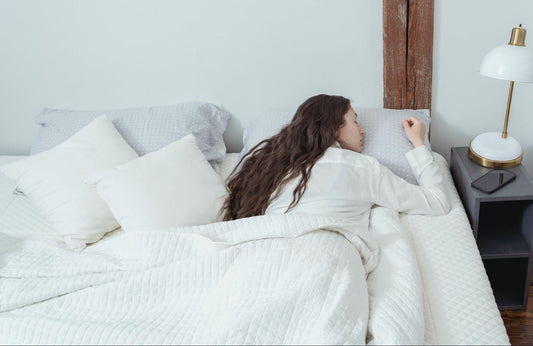 Woman lying on a memory foam pillow on a white bed, under the blanket, with her phone and lamp on the nightstand
