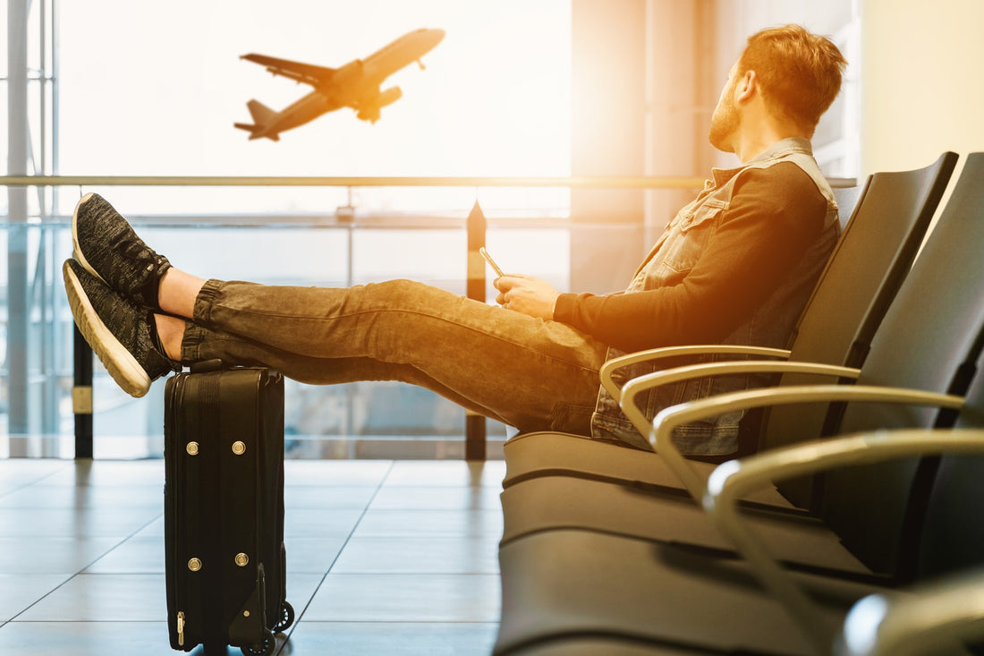 A man sitting at an airport terminal with his legs on his suitcase watching a plane take off outside